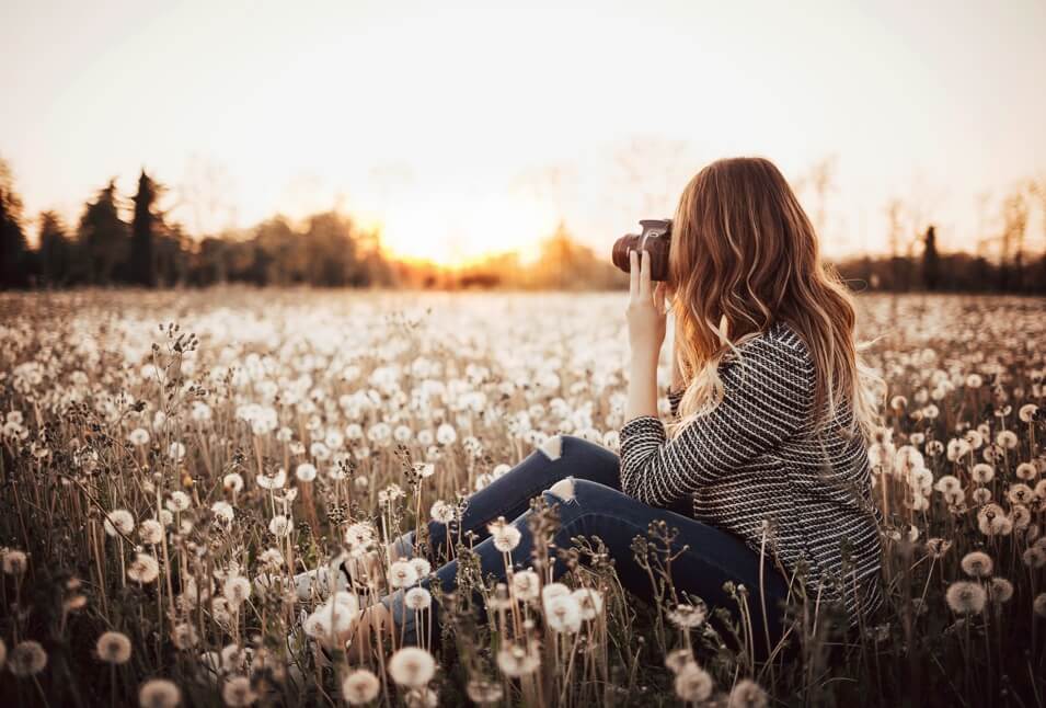 A lady sitting on a beautiful flower field and taking picture of the landscape around her.